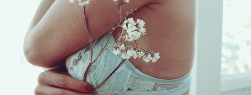 autopalpazione, cancro al seno: busto di una donna di profilo che indossa un reggiseno bianco e afferra un ramo di fiori bianchi