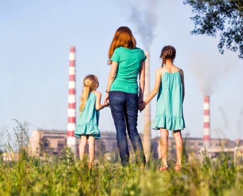 salute e aria inquinata: una donna di spalle mentre tiene per mano due bambine, all'orizzonte i fumi di una fabbrica