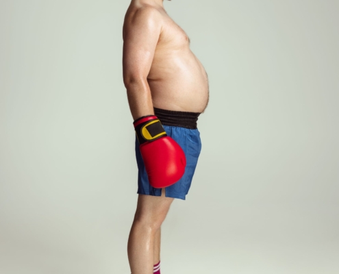obesità: un uomo di profilo con capelli rossi e pancia scoperta che sporge in vista indossa guantoni rossi da box e pantaloncini blue