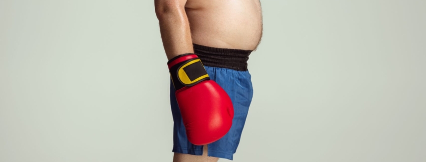 obesità: un uomo di profilo con capelli rossi e pancia scoperta che sporge in vista indossa guantoni rossi da box e pantaloncini blue