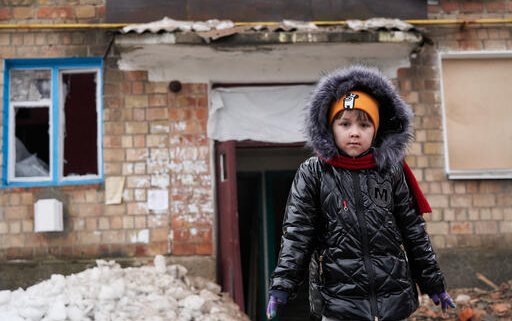 giornata mondiale infanzia, una bimba con alle spalle la casa distrutta dalla guerra in ucraina